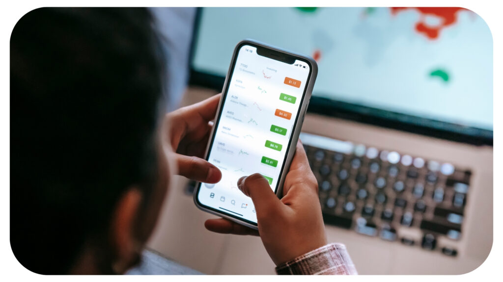 person using trading app on smartphone against laptop.