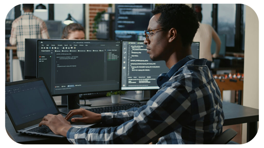 Focused programer fixing glasses while typing on laptop sitting at desk with multiple screens parsing code.