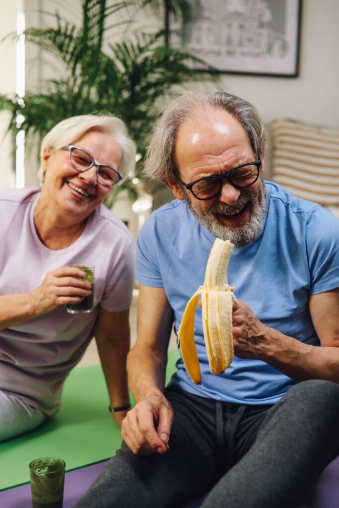 Older couple laughing together at home while enjoying a smoothie and a banana.