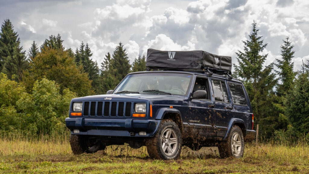 Dark blue Jeep Cherokee parked on a grassy trail with a roof-mounted camping tent, surrounded by trees and cloudy skies.