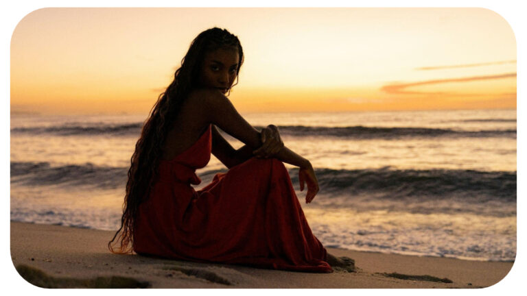Woman wearing a flowing red dress sitting on a sandy beach at sunset, looking over her shoulder as gentle ocean waves roll onto the shore.