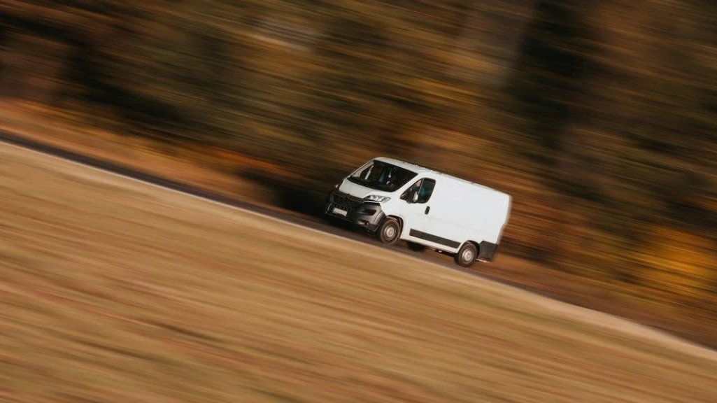 White commercial delivery van driving along a road, with the surrounding landscape blurred by motion.