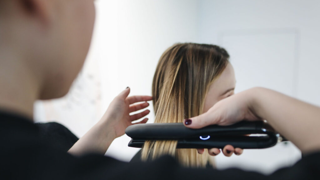 Hair stylist using a flat iron to straighten a woman's blonde hair during a professional styling session.
